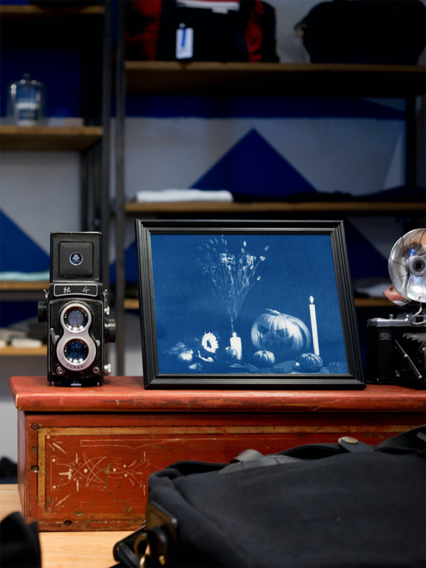 Photographie cyanotype mise en situation dans un cadre noir. Verso de ma nature morte qui représente une grosse citrouille avec les yeux et le sourire façon Halloween, ainsi que deux autres mini citrouilles, des bougies allumées, un bouquet de fleurs séchées, un tournesol, des pommes et des feuilles mortes.
