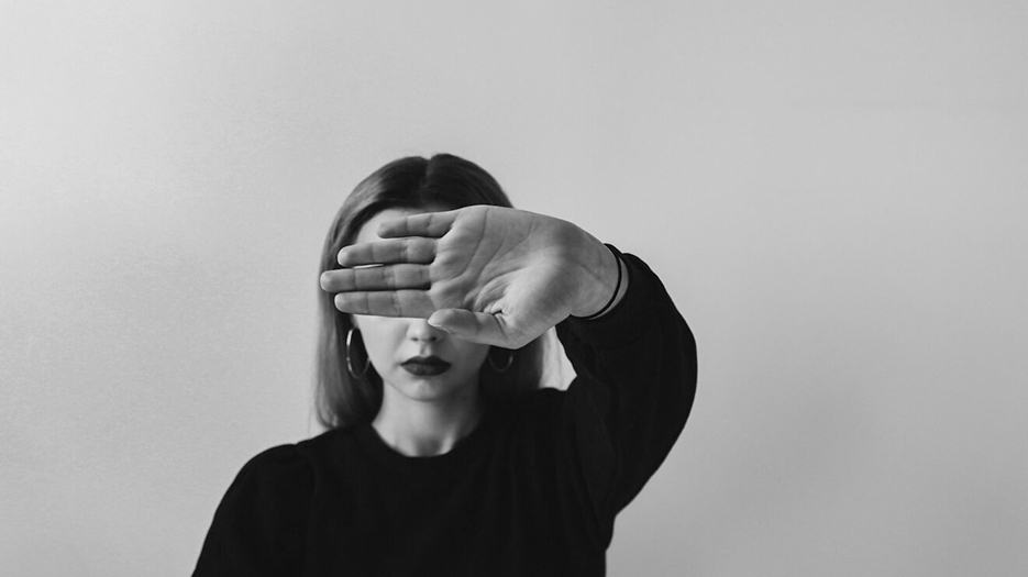 Photo en noir et blanc d'une jeune femme blanche qui tend sa main en avant et sur laquelle la mise au point est faite. Ainsi, la main, paume vers l'objectif, cache les yeux de la jeune femme.
