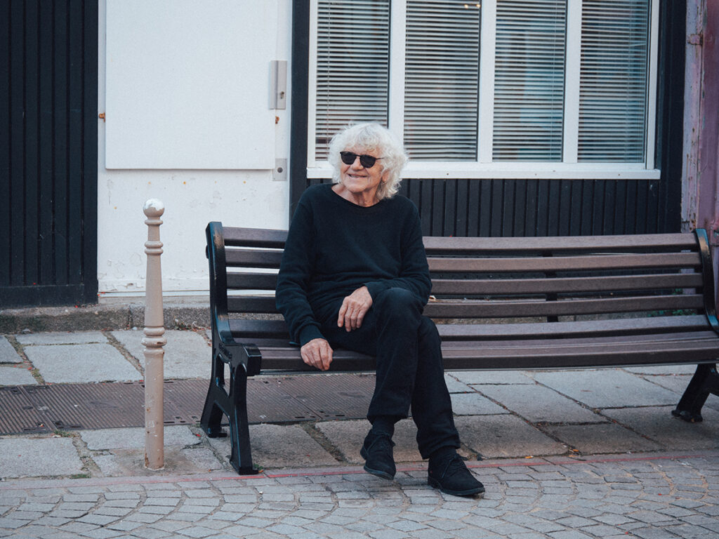 Personne âgée vêtue de noir et avec des lunettes de soleil, assise sur un banc et faisant un large sourire.