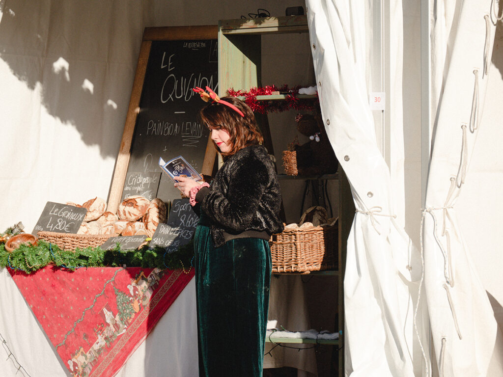 Jeune femme en train de lire un prospectus. Elle porte une combinaison en velours vert, un blouson noir à paillettes et un serre-tête rouge qui représente des bois de renne. Elle est face à son stand du marché de Noël, stand sur lequel elle vend du pain.