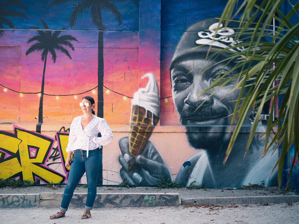 Femme avec des lunettes de soleil miroir souriante devant une fresque qui représente Snoop Dogg en train de manger une glace.