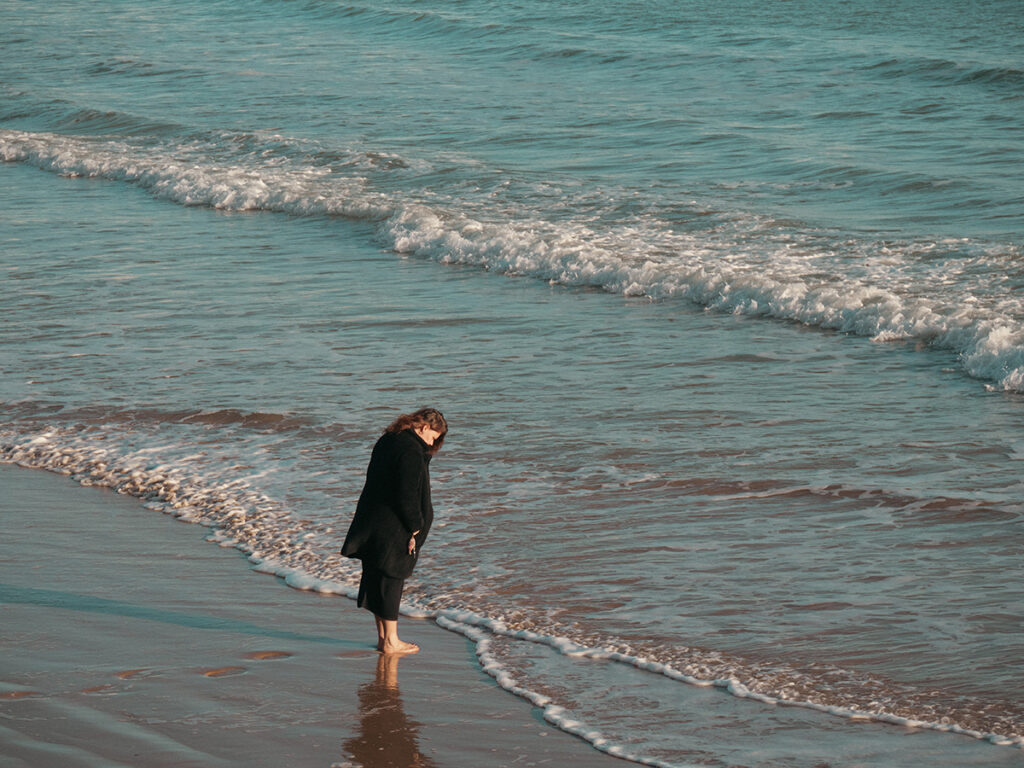 Femme habillée en noir qui regarde l'océan, tête baissée, les pieds dans l'eau.