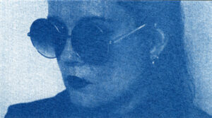 Autoportrait monochrome bleu, plan très serré sur mon visage. Je porte des lunettes de soleil rondes et mon visage est disposé de 3/4.