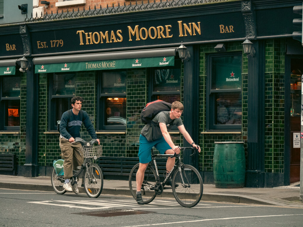 Deux jeunes hommes blancs à vélo, arrêtés au feu. Ils sont devant un pub, le Thomas Moore Inn.