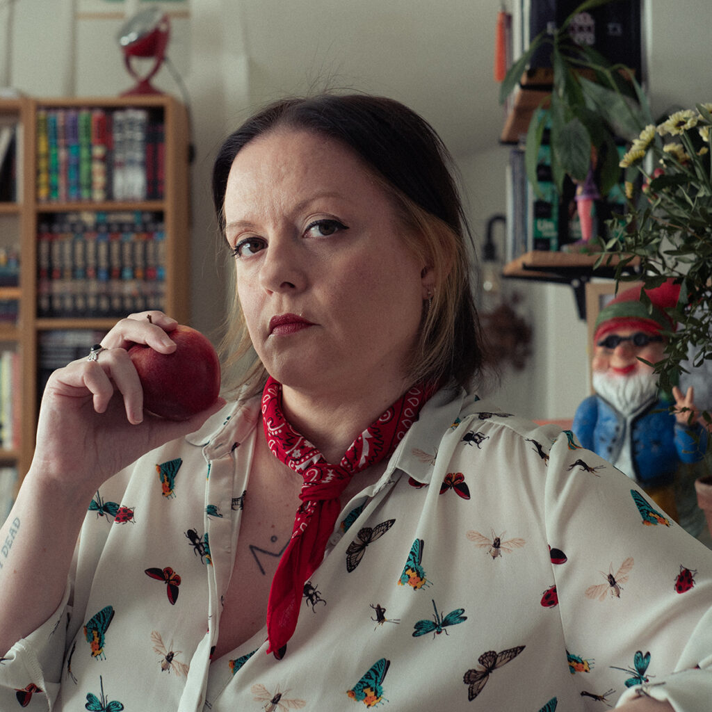 Autoportrait à la façon Blanche-Neige. Je porte mes cheveux en carré court, ils sont bruns avec quelques mèches blondes. Je porte une chemise avec un imprimé insectes, un bandana rouge autour du cou et une pomme rouge dans la main. En arrière-plan, mon nain de jardin.