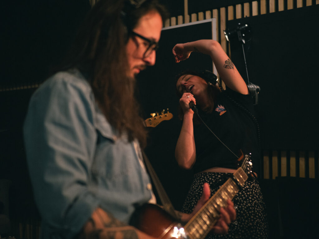 Le guitariste et la chanteuse du groupe Amante. Le guitariste a les cheveux longs et des lunettes à monture épaisse. La chanteuse rejette la tête et le bras en arrière.