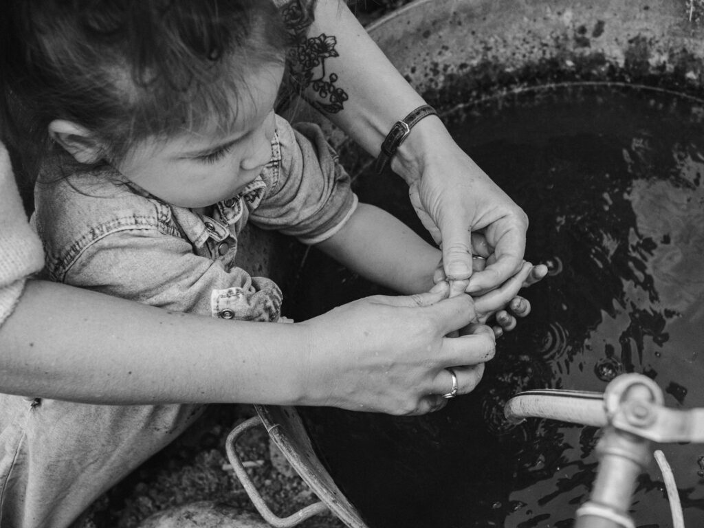 Photo en noir et blanc d'une petite fille qui se lave les mains dans une cuve transformée en évier. La petite est aidée par sa maman dont on ne voit que les bras.
