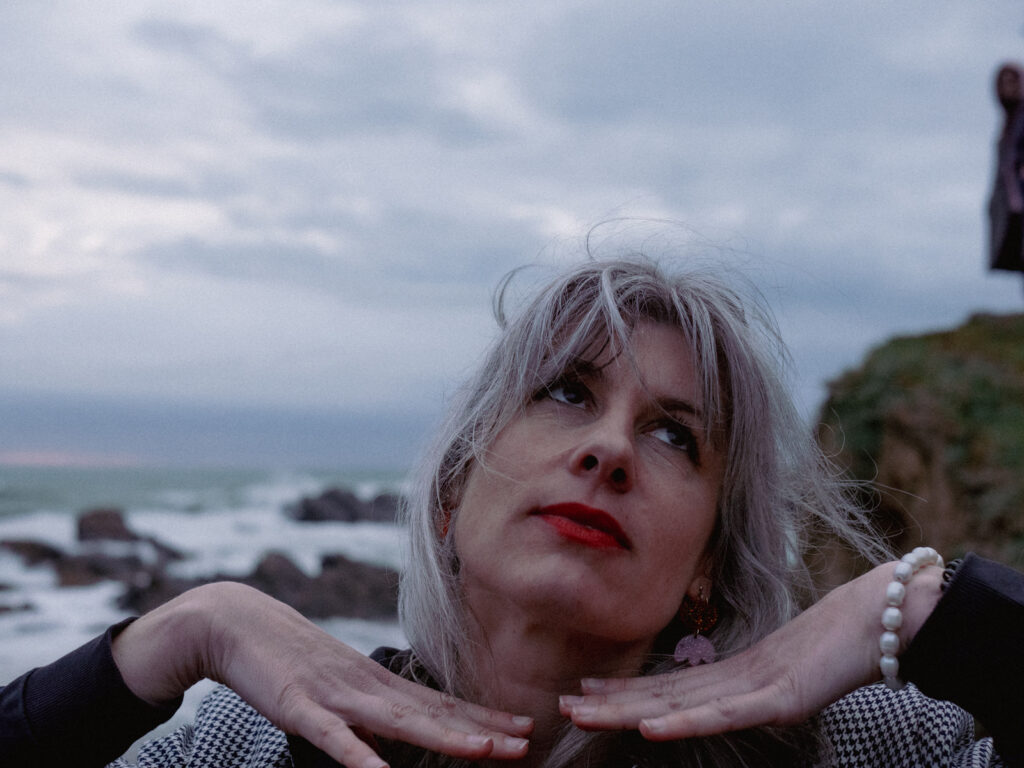 Portrait d'une femme fraîchement quarantenaire. Elle est blanche, ses cheveux aussi, et elle pose devant l'océan d'avril, les mains sous le menton et le regard au loin, la frange au vent.