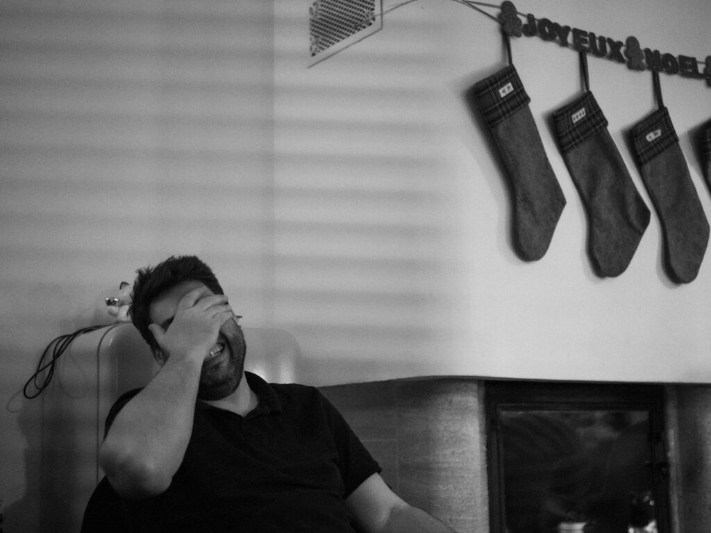 Portrait en noir et blanc d'un homme qui rit avec la main flanquée sur ses yeux. Il est assis dans un fauteuil et on voit les chaussettes de Noël accrochées au-dessus de la cheminée.