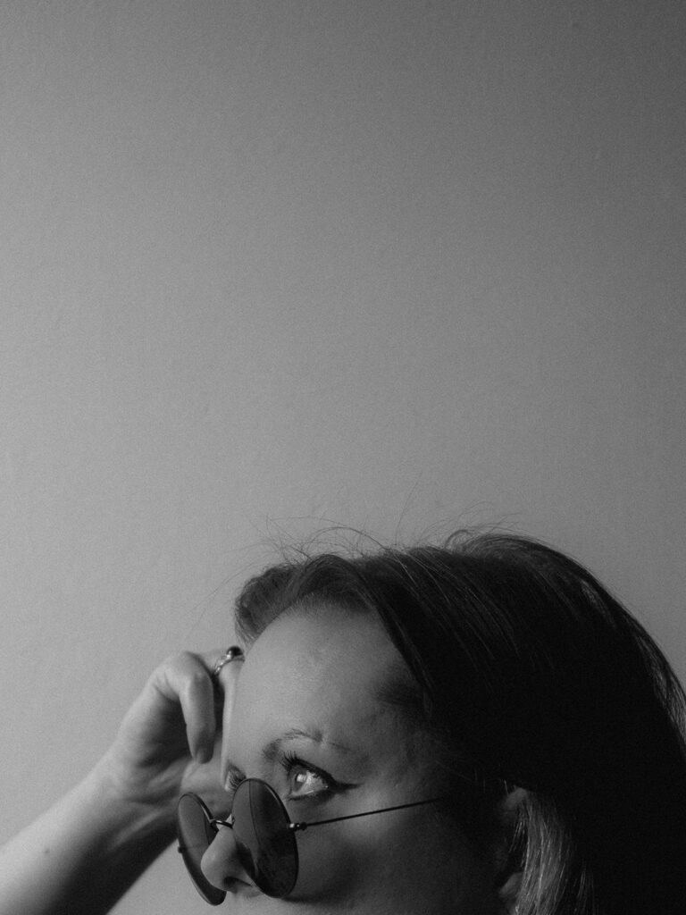 Autoportrait en noir et blanc. Profil gauche coupé sous le niveau du nez. Lunettes de soleil rondes, eyeliner, regard porté en l'air. Main droite posée sur la tempe.