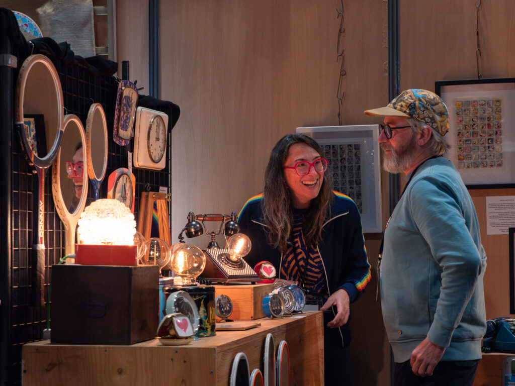 Une artisane sur son stand d'objets upcyclés, notamment des horloges, tout sourire avec un client curieux.