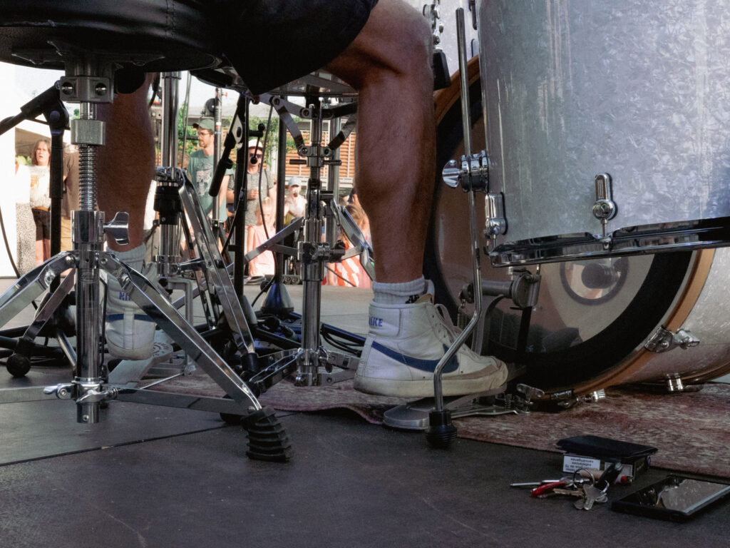Photo centrée sur les pieds d'un batteur en short et en basket, on voit ses clefs, son portefeuille, son smartphone et son paquet de clopes posés par terre, cachés aux yeux du public.