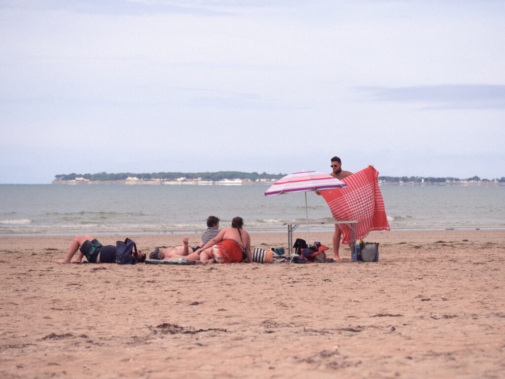 Une famille installée sur la plage, à Noirmoutier. Tout le monde est assis, sauf un homme qui déplie une grande serviette.