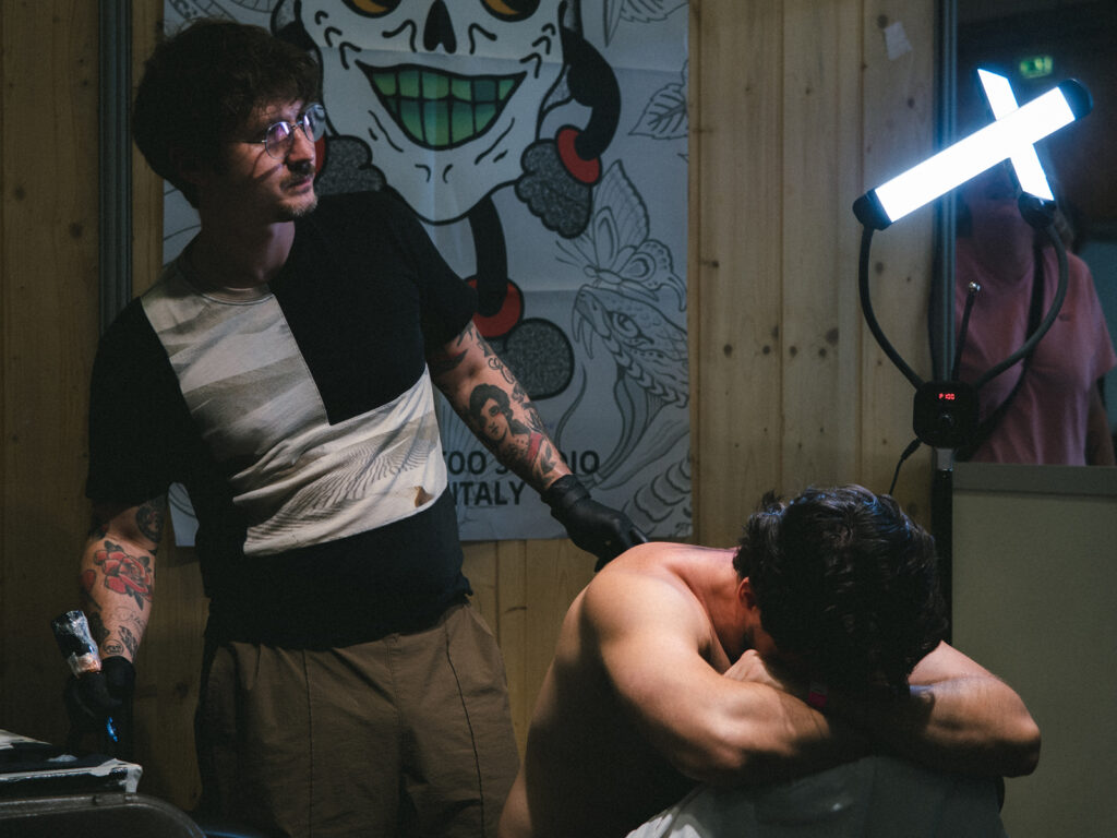Un tatoueur s'apprête à tatouer un homme torse nu, assis à cheval sur une chaise et la tête reposant sur les bras. Les deux hommes sont éclairés par une lampe d'appoint en forme de croix chrétienne.