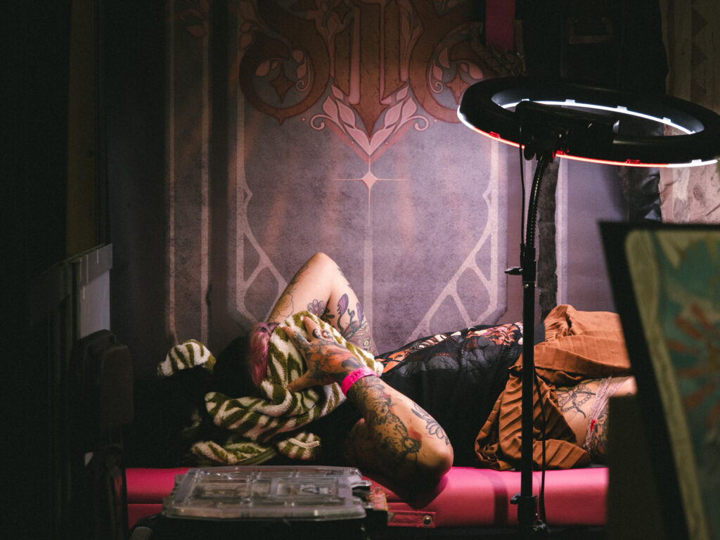 Une femme en train de se faire tatouer, le visage caché par un vêtement imprimé, elle semble souffrir. La lumière est rosée et tamisée.