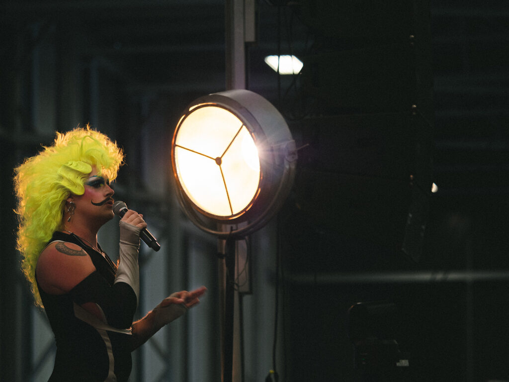 La drag queen Jodye Coaster sur scène avec un micro et une perruque jaune fluo, face à un grand projecteur.