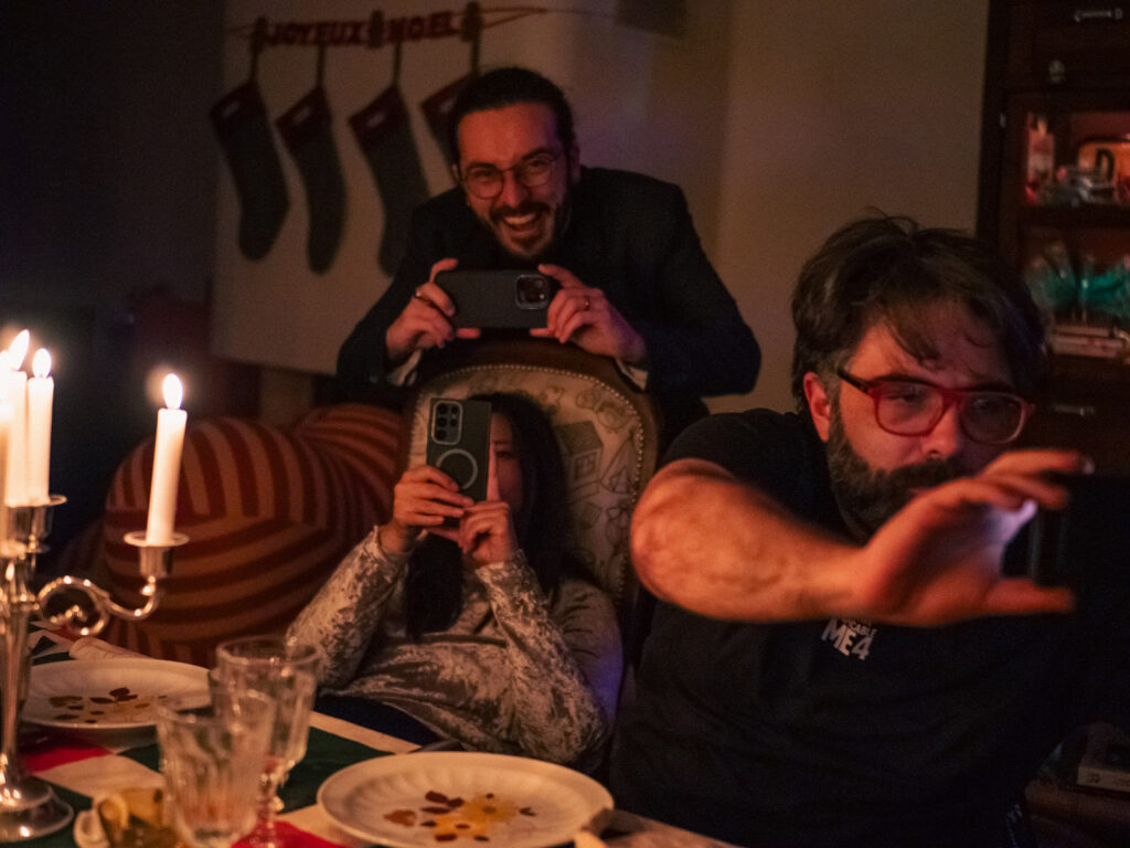 Un groupe d'amis à une table de réveillon, tout le monde braque son smartphone vers la photographe.