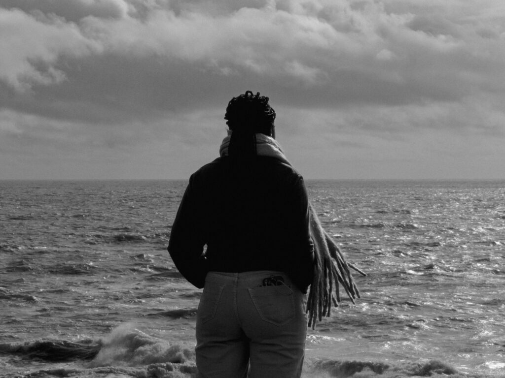 Photo en noir et blanc d'une jeune femme noire, de dos, qui se tient face à l'océan.