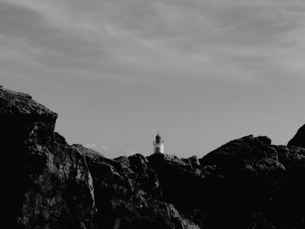 Photo en noir et blanc de sommet d'un phare qui surgit de derrière les rochers.