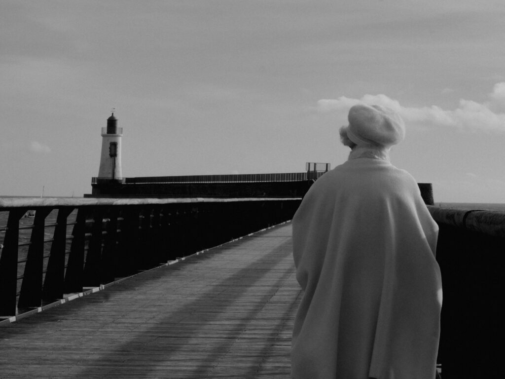 Photo en noir et blanc d'une vieille dame vue de dos, elle porte une cape et un béret blancs et se dirige vers le phare, au loin, via la jetée.
